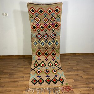 May include: A rectangular, beige-colored rug with a geometric pattern of diamonds and crosses in red, black, green, blue, and pink. The rug has a fringe on the bottom edge and is displayed on a wooden floor.