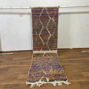 Puede incluir: Una alfombra de pasillo marroquí colorida con un patrón de diamantes en tonos de rosa, morado, verde y amarillo. La alfombra está hecha de lana y tiene flecos en ambos extremos.