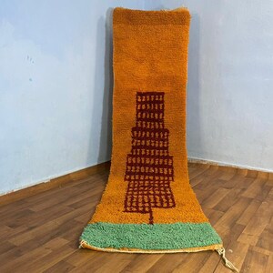 May include: A long, rectangular rug with a geometric design. The rug is orange with a brown and red geometric pattern in the centre. The bottom of the rug is green.