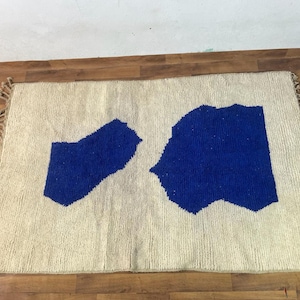 May include: A white rug with two blue abstract shapes. The rug has fringe on all sides.