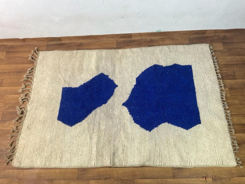 May include: A white rug with two blue abstract shapes. The rug has fringe on all sides.