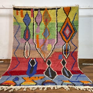 May include: A colorful Moroccan rug with geometric patterns in shades of red, blue, green, yellow, and orange. The rug has a fringe border and is woven with a thick, textured yarn.
