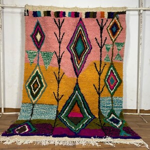May include: A colorful Moroccan rug with a geometric pattern in shades of pink, yellow, green, blue, and purple. The rug has a fringe border and is made of wool.