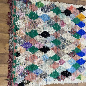 Puede incluir: Una alfombra marroqu&iacute; colorida con un patr&oacute;n de diamantes en tonos de verde, azul, rosa y negro. La alfombra tiene un fondo blanco y est&aacute; hecha de lana.