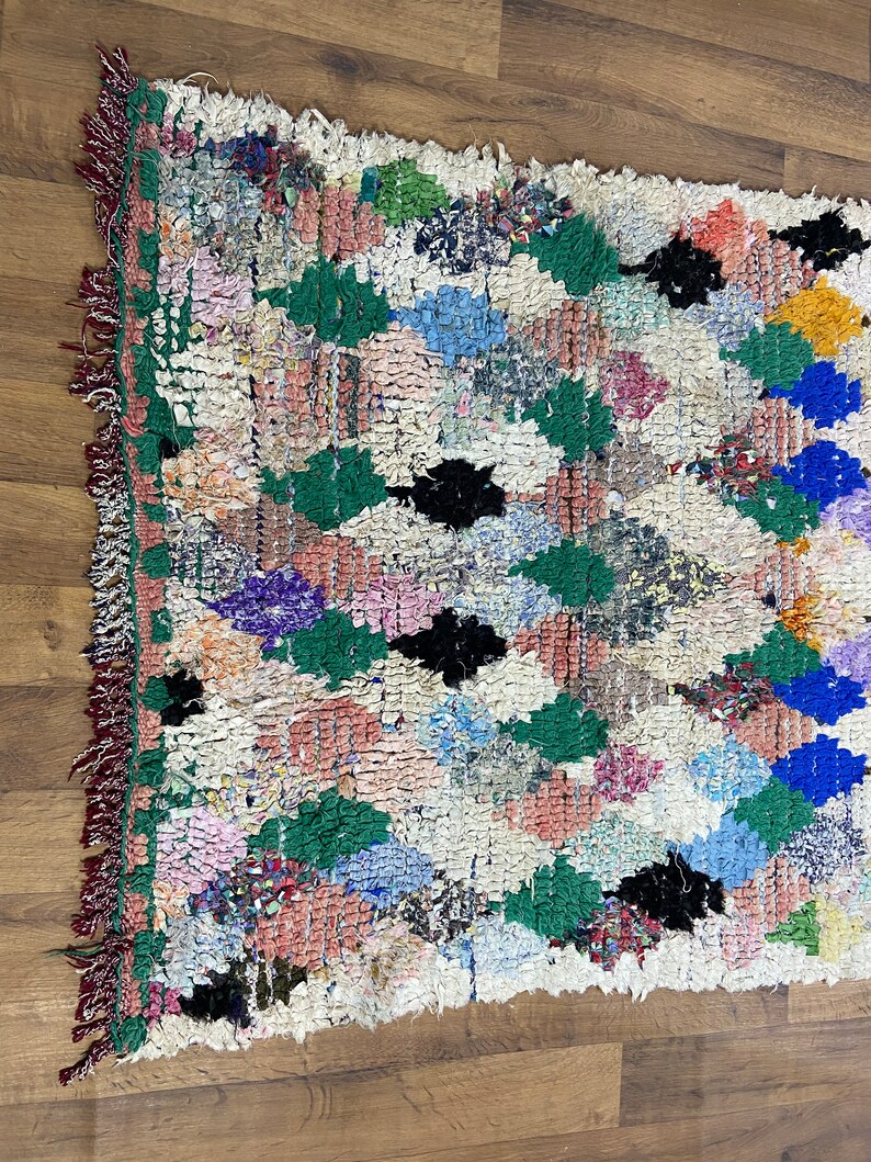 Puede incluir: Una alfombra marroqu&iacute; colorida con un patr&oacute;n de diamantes en tonos de verde, azul, rosa y negro. La alfombra tiene un fondo blanco y est&aacute; hecha de lana.