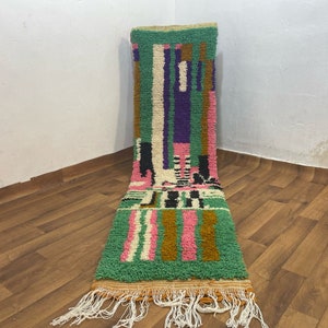 May include: A long, narrow rug with a geometric pattern in shades of green, pink, white, black, and brown. The rug has a fringe on both ends.