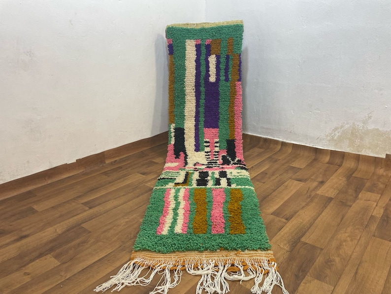May include: A long, narrow rug with a geometric pattern in shades of green, pink, white, black, and brown. The rug has a fringe on both ends.