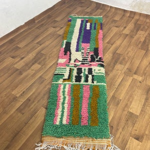 May include: A long, rectangular rug with a colourful geometric pattern. The rug is made of wool and has a fringe on the ends. The colours in the rug are green, pink, brown, white, and black.