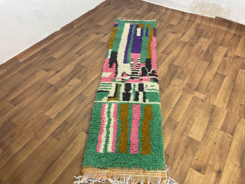 May include: A long, rectangular rug with a colourful geometric pattern. The rug is made of wool and has a fringe on the ends. The colours in the rug are green, pink, brown, white, and black.