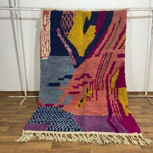 May include: A colorful, abstract rug with a geometric design. The rug features a mix of colors including blue, pink, yellow, and purple. It has a fringed edge and is displayed on a white rack.