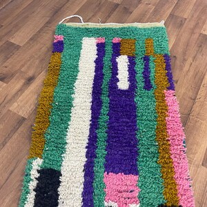 May include: A colourful, handwoven rug with geometric patterns in shades of green, purple, pink, white, and brown. The rug has a thick, textured pile and is made with natural fibres.