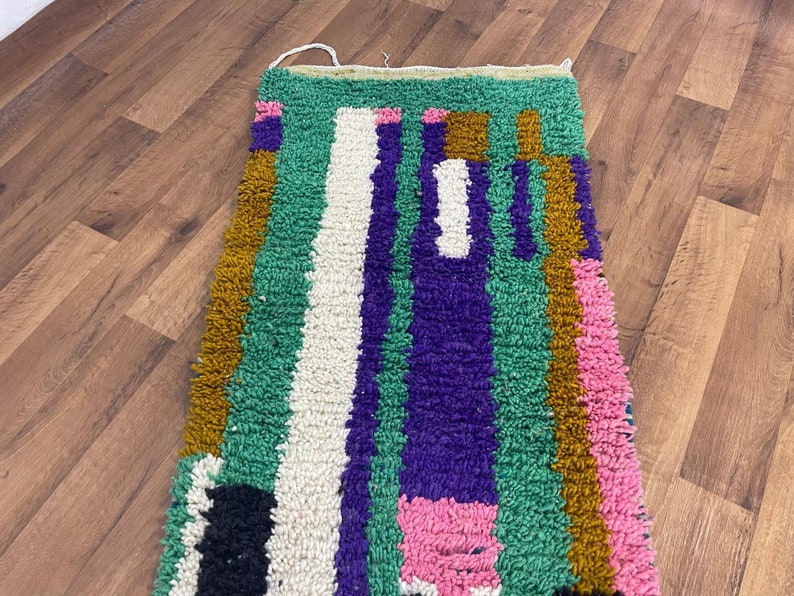 May include: A colourful, handwoven rug with geometric patterns in shades of green, purple, pink, white, and brown. The rug has a thick, textured pile and is made with natural fibres.