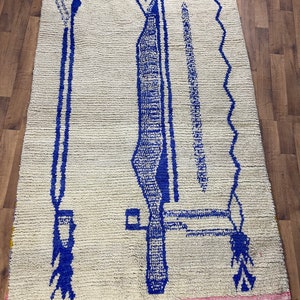 May include: A white Moroccan rug with a blue geometric design. The rug has a pink fringe around the edges.