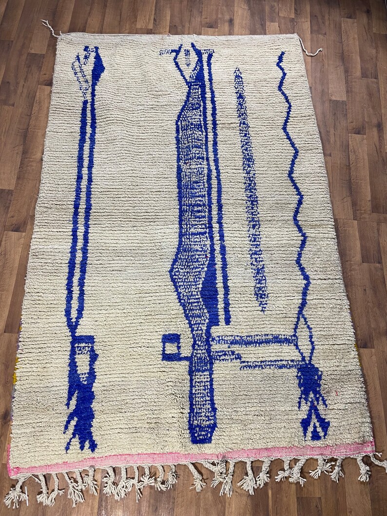 May include: A white Moroccan rug with a blue geometric design. The rug has a pink fringe around the edges.