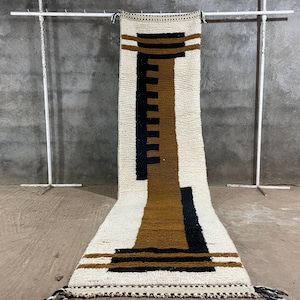 Könnte beinhalten: Ein rechteckiger, cremefarbener Teppich mit einem geometrischen Design in Schwarz und Braun. Der Teppich wird vertikal präsentiert, mit einem sich wiederholenden Muster aus Linien und Formen. Der Teppich hat einen Fransen am unteren Rand und wird auf einem Metallgestell ausgestellt.