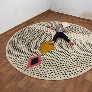 Può includere: Un grande tappeto rotondo con un motivo geometrico di punti neri e spazi bianchi. Presenta accenti a diamante gialli e rosa. Il tappeto è su un pavimento in legno.