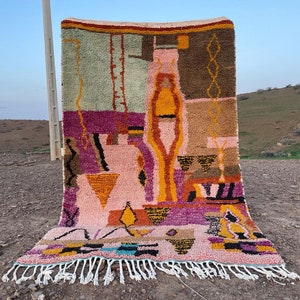 May include: A colorful Moroccan rug with geometric patterns in shades of pink, orange, yellow, brown, and green. The rug has a fringe border and is displayed outdoors.