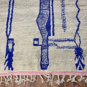 May include: A white rug with a blue geometric pattern. The rug has a fringe border.