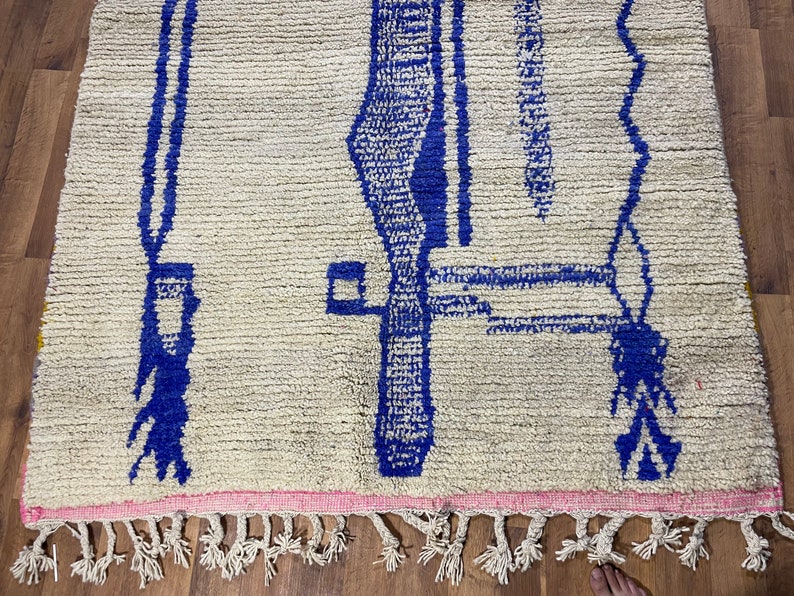 May include: A white rug with a blue geometric pattern. The rug has a fringe border.