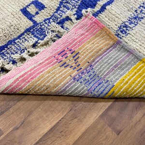 May include: A close-up of a colourful woven rug with a fringe. The rug has stripes of pink, yellow, blue, and purple. The rug is folded in half, revealing the underside.