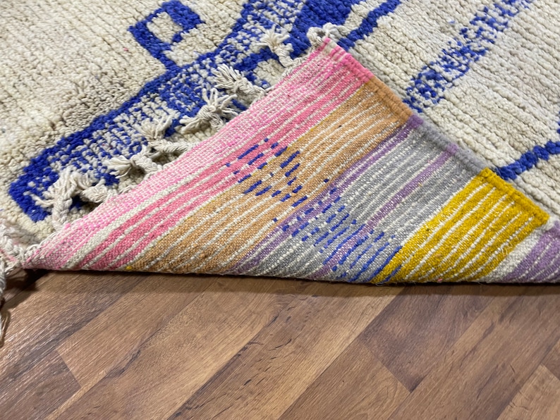 May include: A close-up of a colourful woven rug with a fringe. The rug has stripes of pink, yellow, blue, and purple. The rug is folded in half, revealing the underside.