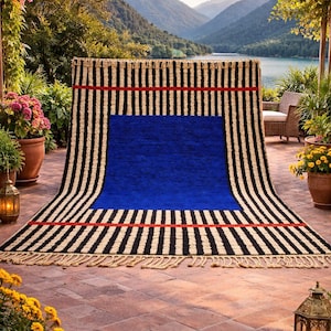 May include: A rectangular area rug featuring a large central solid blue square surrounded by vertical black and cream stripes. Two horizontal red bands cross the striped sections, and the top and bottom edges have decorative fringe.