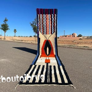 Puede incluir: Una alfombra larga y estrecha, colorida, con un patrón geométrico. La alfombra está hecha de lana y tiene flecos en los extremos. La alfombra está colocada sobre una superficie pavimentada.