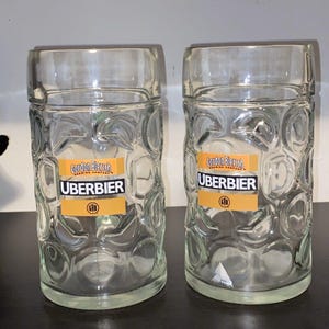 May include: Two clear glass beer steins with a textured, dimpled design. Each stein has a rectangular label with the words "Gordon Biersch Brewing Company" and "UBERBIER" printed in black. The steins are for beer.