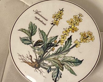 May include: A white ceramic plate with a brown rim features a botanical illustration of a yellow flower with green leaves and the text "Agrimonia eupatoria".