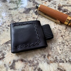 May include: A black leather pen holder with a stitched border and an elastic band. A wooden pen with gold accents rests nearby. The pen holder is designed to attach to a notebook or journal, providing a convenient place to store a pen.