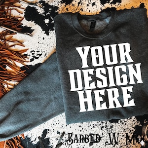 May include: A dark grey sweatshirt with the text "YOUR DESIGN HERE" in white, displayed on a cowhide print surface. The sweatshirt is a mock-up for custom designs. The image also includes leather and metal accessories.