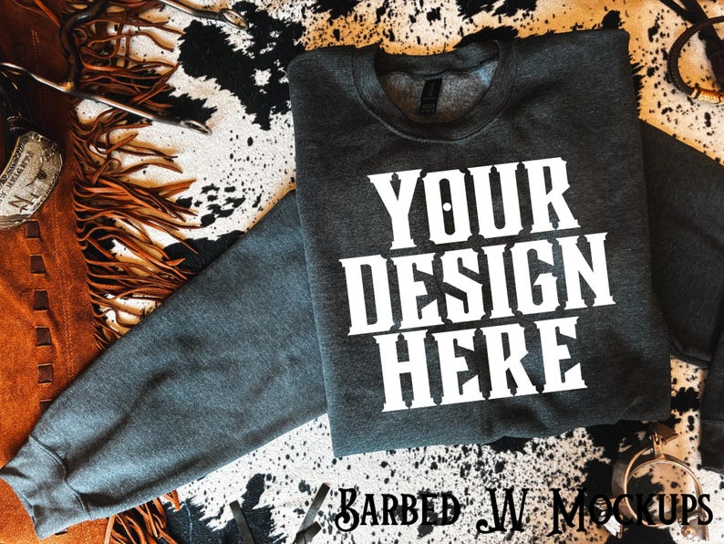 May include: A dark grey sweatshirt with the text "YOUR DESIGN HERE" in white, displayed on a cowhide print surface. The sweatshirt is a mock-up for custom designs. The image also includes leather and metal accessories.
