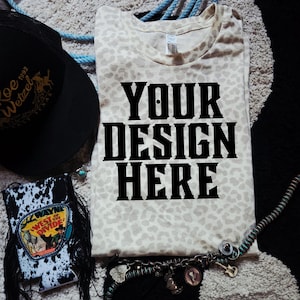May include: A cream-colored t-shirt with a leopard print pattern and the text "YOUR DESIGN HERE" in bold black letters. A black baseball cap, a drink holder, and a charm bracelet are also visible.