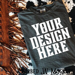 May include: Dark gray t-shirt with the text "YOUR DESIGN HERE" in white, displayed on a cowhide-patterned surface. A brown leather item with fringe is on the left side of the image. The shirt has a Comfort Colors tag.