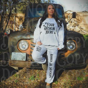 Może przedstawiać: Jasnoszary zestaw bluzy i spodni dresowych. Bluza i spodnie dresowe mają nadruk "Your Design Here". Spodnie dresowe mają również nadruk "Barbed JW Mockups" z boku. Modelka ma na sobie szachownicowe wsuwane buty.