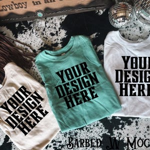 May include: Three folded t-shirts in beige, teal, and white, each with the text "YOUR DESIGN HERE" in a bold, black font. The shirts are arranged on a cowhide-patterned surface, with decorative elements in the background.