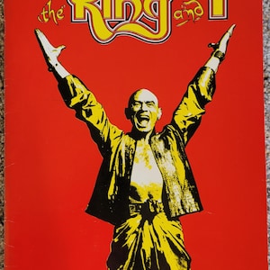 Op de afbeelding: Een poster voor de musical "The King and I". De poster toont een kale man met opgeheven armen tegen een rode achtergrond. De man draagt een gele outfit. De titel staat in gele en zwarte tekst.