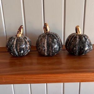 May include: Three ceramic pumpkins with a dark grey and brown speckled finish. Each pumpkin has a gold-coloured stem. The pumpkins are displayed on a wooden shelf against a white background.