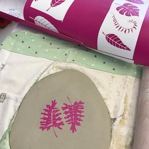 May include: A rolled magenta and white stencil featuring leaf patterns is positioned above a grey clay piece with two magenta leaf imprints. The clay rests on a green and white polka dot cloth.
