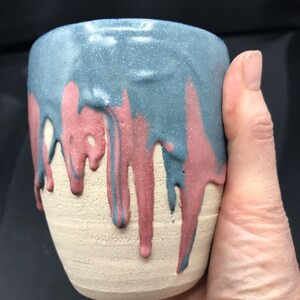 May include: A handcrafted ceramic cup with a distinctive design. The upper section is speckled blue, with red and blue drips flowing down to the unglazed, beige base. The cup is held in a hand, highlighting its form and size.