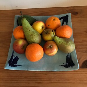 May include: A light blue square ceramic platter filled with fresh fruit. The fruit includes oranges, apples, and pears. The platter has dark blue brushstroke designs on the corners. The fruit is arranged on a wooden surface.