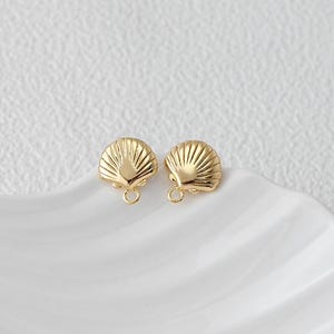 May include: Two gold-colored seashell charms with a loop for jewelry making. The charms are fan-shaped with detailed ridges, and are displayed on a white, wavy surface. The charms are small and delicate.