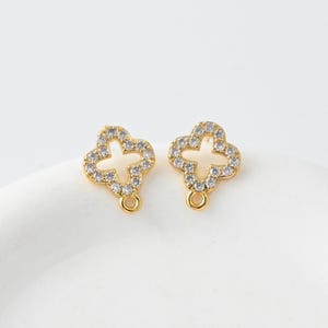 Peut inclure: Deux breloques dorées en forme de trèfle à quatre feuilles, ornées de cristaux clairs et étincelants. Chaque breloque possède une petite boucle en bas pour l'attache de bijoux. Les breloques sont présentées sur une surface blanche.