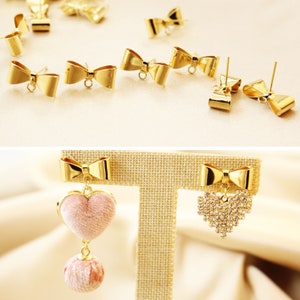 Puede incluir: Abalorios de pendientes de lazo dorados con un dije en forma de corazón y un dije en forma de corazón de pedrería. Los dijes cuelgan de los abalorios de lazo.
