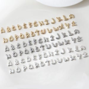 May include: A selection of gold and silver alphabet charms. The charms are in a bubble letter style, with each letter of the alphabet represented. The charms are small and have a loop at the top for attaching to jewellery or other crafts.
