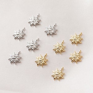 Puede incluir: Diez colgantes de sol de oro y plata. Los colgantes están detallados con una cara y rayos. Los colgantes están dispuestos sobre una superficie blanca.