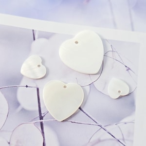 May include: Four white heart-shaped shell pendants with holes for stringing. The pendants are different sizes and are arranged on a white surface.