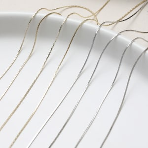 May include: A close-up of six silver and gold chains on a white surface. The chains are all the same length and have a delicate, textured design.