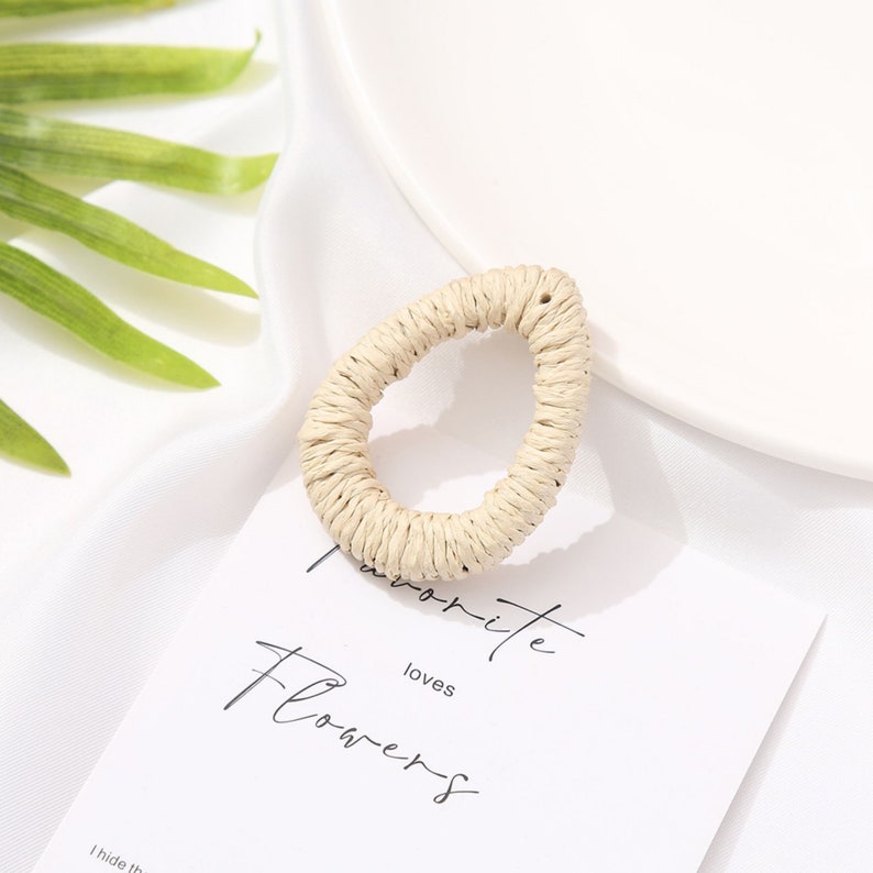 May include: A teardrop-shaped earring made of woven natural fibers. The earring is a light beige color and has a textured surface. The earring is on a white background with a card that says "Favorite Flowers".