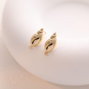 May include: Two gold-coloured seashell-shaped earring components. Each component has a spiral top and a rounded, oval body with a loop at the bottom. The earrings are on a white surface.